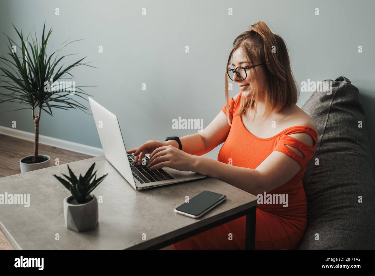 Caucasian Young Woman in the Red Dress Working by Laptop from Home, Female Freelancer at Work, Remote Job Stock Photo