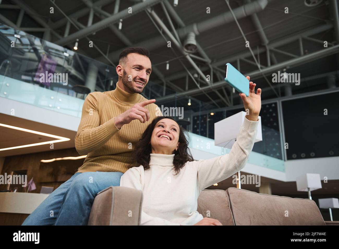 Handsome young multi ethnic couple in casual wear holding smartphone and  looking at webcam talking via video call while relaxing in vip lounge  waiting Stock Photo - Alamy