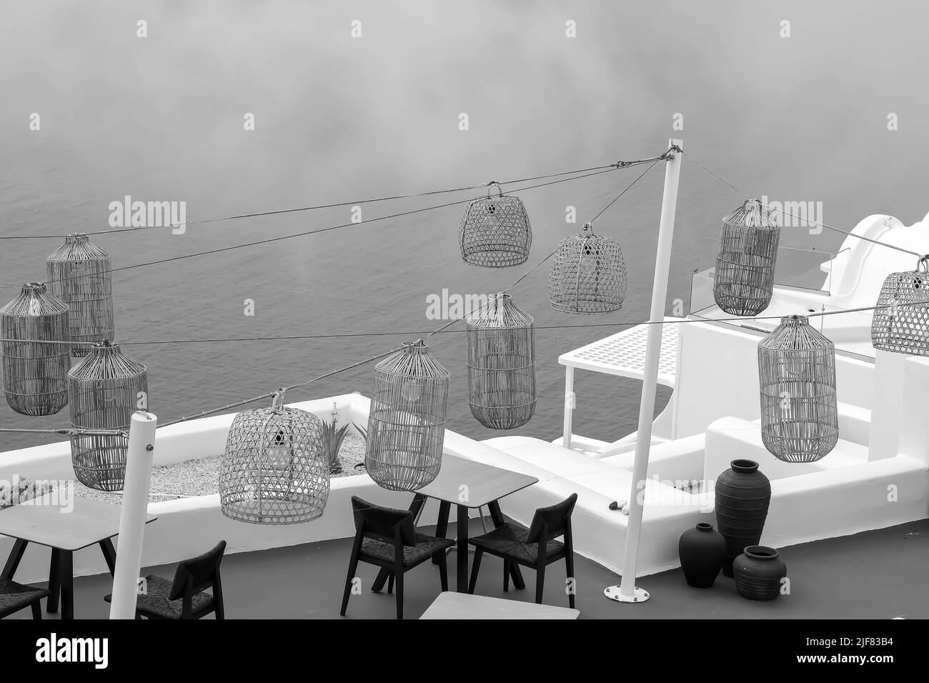 Santorini, Greece - May 13, 2021 :  View of a picturesque terrace decorated with tables, chairs  and lights overlooking the Aegean Sea in black and white Stock Photo