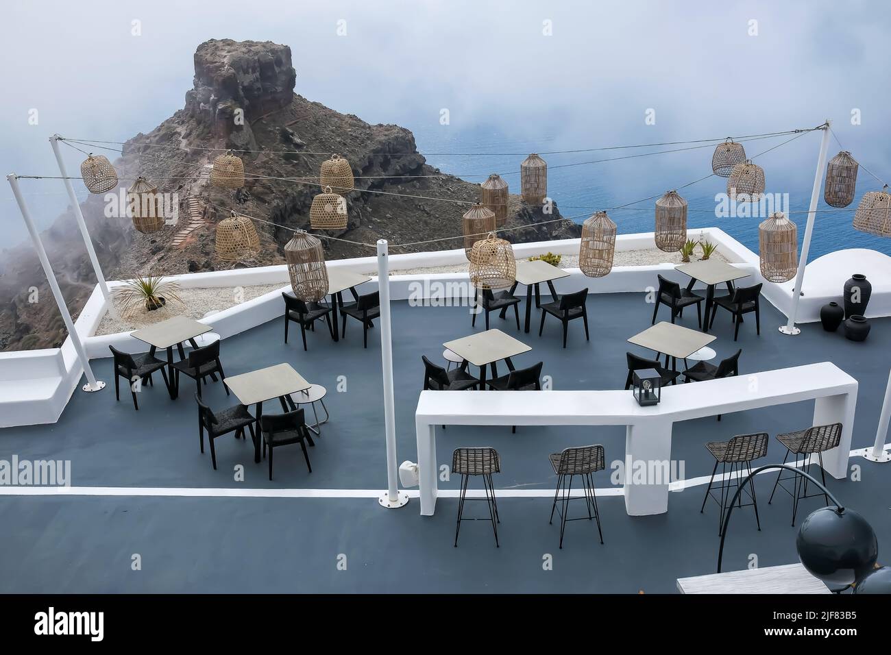 Santorini, Greece - May 13, 2021 :  View of a picturesque terrace decorated with tables, chairs  and lights overlooking a huge rock and the Aegean Sea Stock Photo
