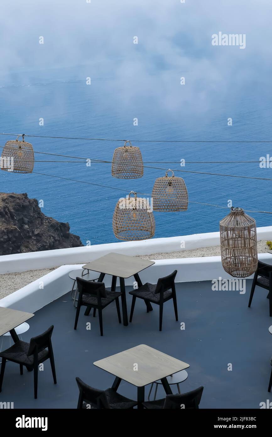 Santorini, Greece - May 13, 2021 :  View of a picturesque terrace decorated with tables, chairs  and lights overlooking a huge rock and the Aegean Sea Stock Photo