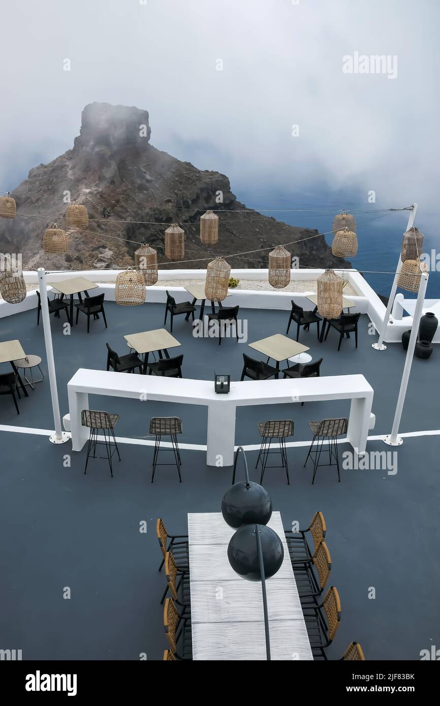 Santorini, Greece - May 13, 2021 :  View of a picturesque terrace decorated with tables, chairs  and lights overlooking a huge rock and the Aegean Sea Stock Photo
