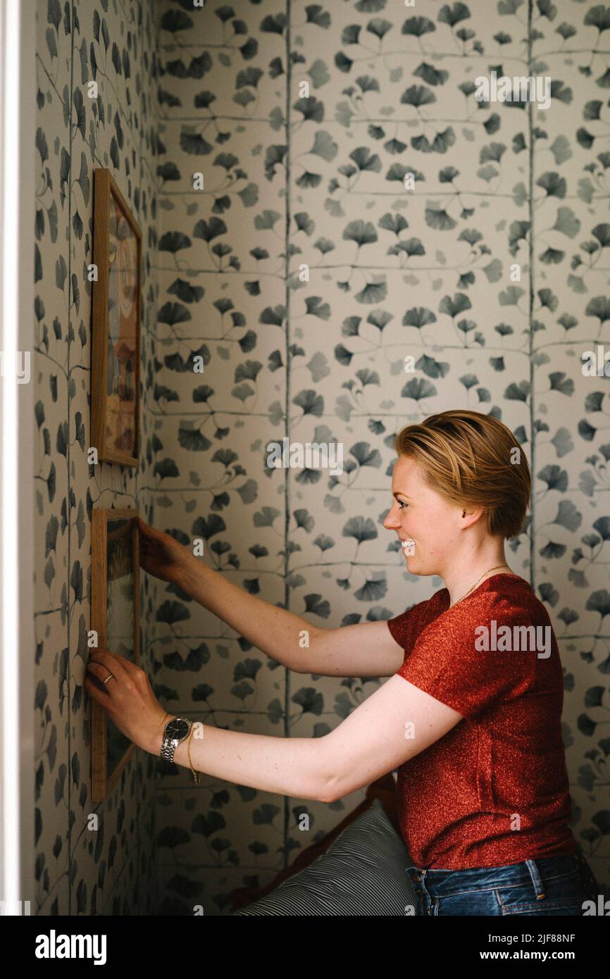 Side view of smiling woman adjusting picture frame on wall at home Stock Photo