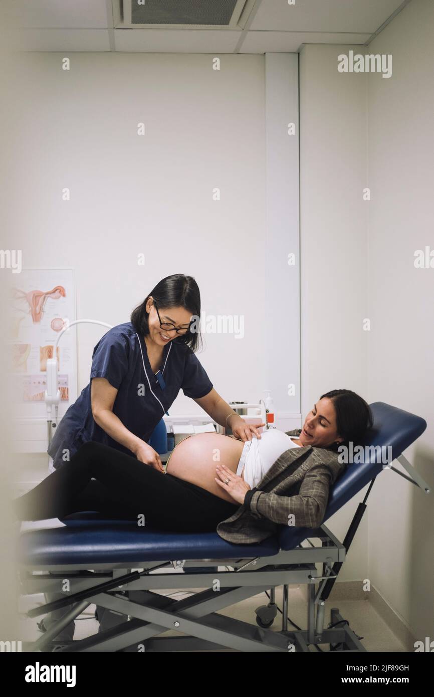 Smiling gynecologist measuring abdomen of pregnant woman lying on gurney in hospital Stock Photo