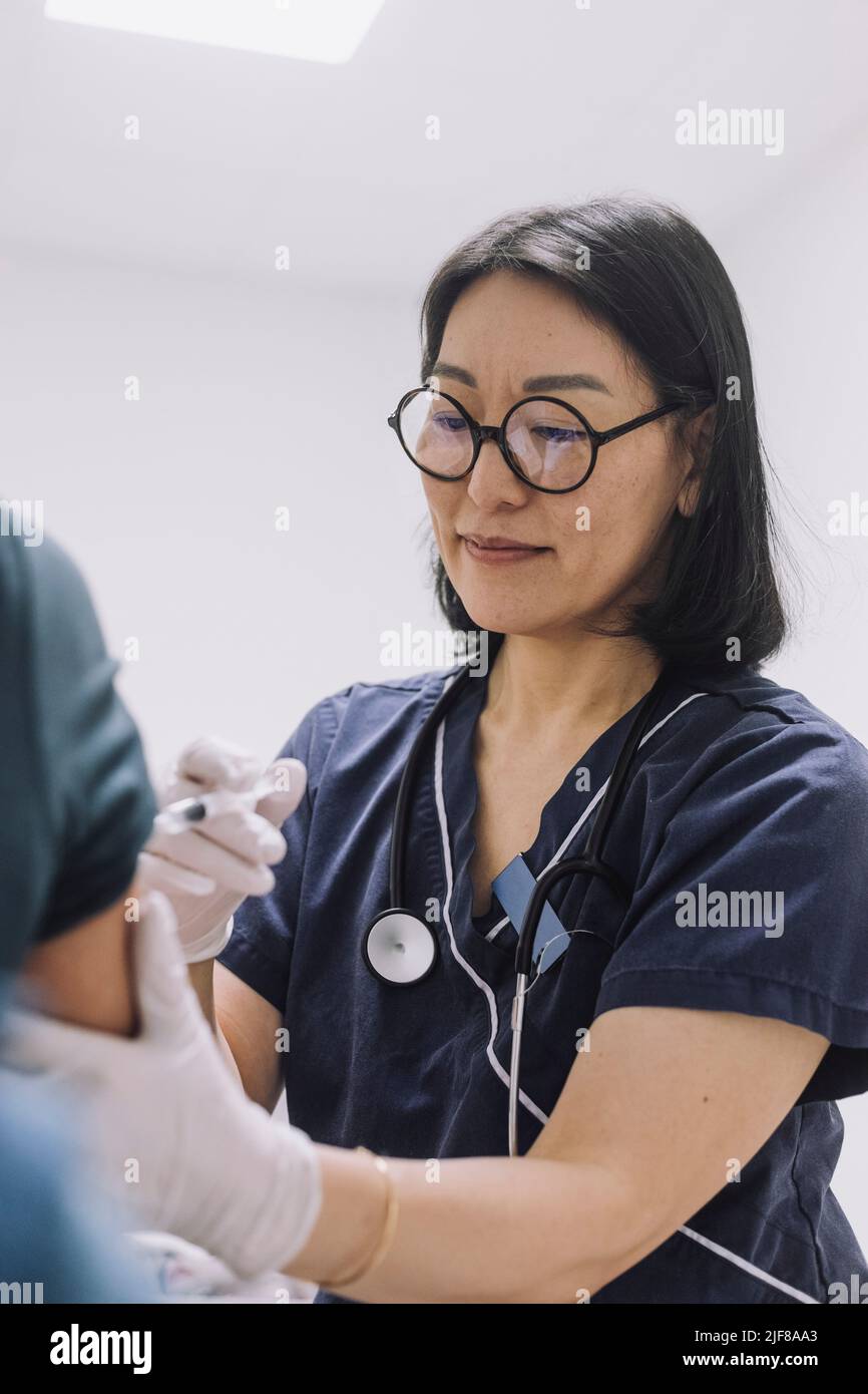 Female doctor wearing eyeglasses injecting patient in hospital Stock