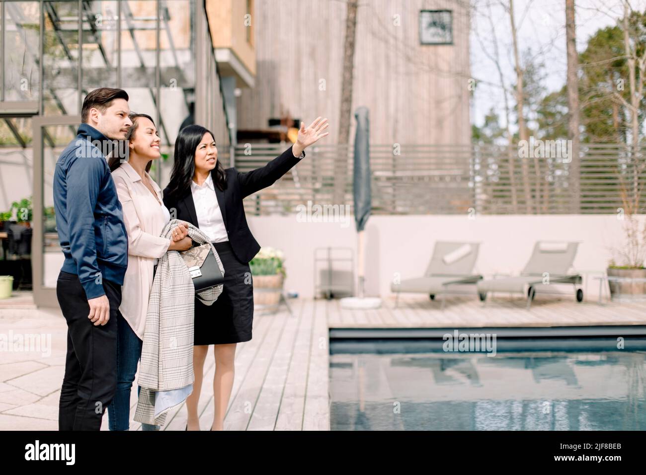 Real estate agent showing backyard to couple while standing at poolside Stock Photo
