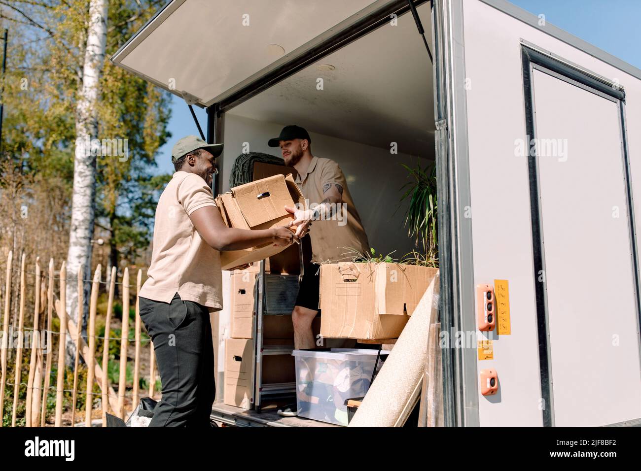 Multiracial movers unloading cardboard boxes from truck during sunny day Stock Photo