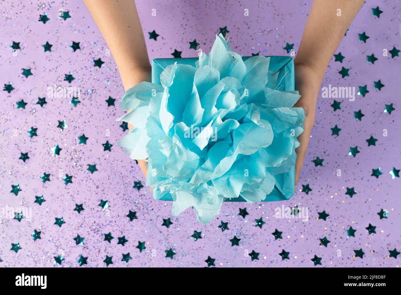 A gift wrapped in blue eco kraft paper with a lush bow on a lilac background with small confetti in the form of stars.The holiday, surprise, birthday. Stock Photo