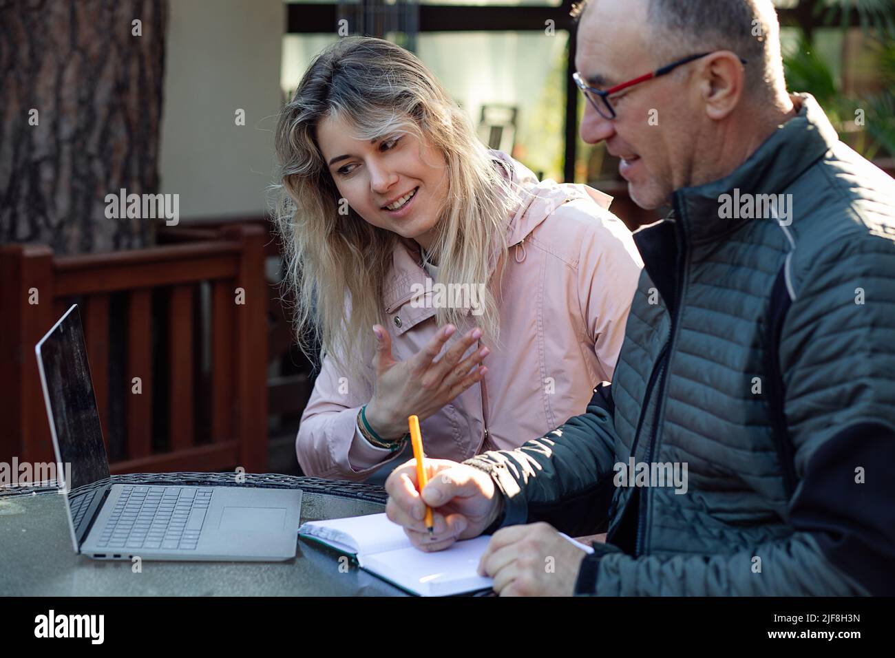 Portrait of young smiling woman daughter trainer talking to middle-aged man, explaining information on screen of laptop. Stock Photo