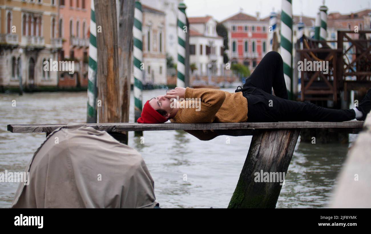 A young man lying on a bench uper the waterfront and smoke cigarettes in  the Venice streets Stock Photo