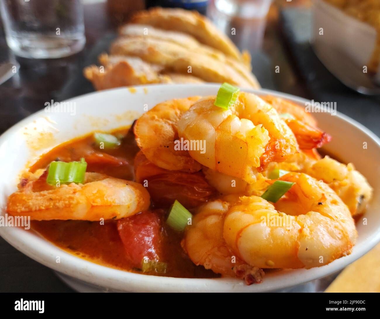 Garlic Shrimp with red sauce and green onions served in a white bowl with bread and a glass of water in the background. Stock Photo