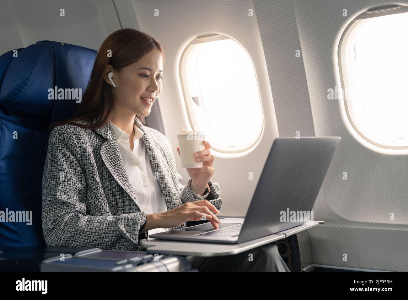 Attractive asian female passenger of airplane sitting in comfortable seat listening music in earphones while working at modern laptop computer with Stock Photo