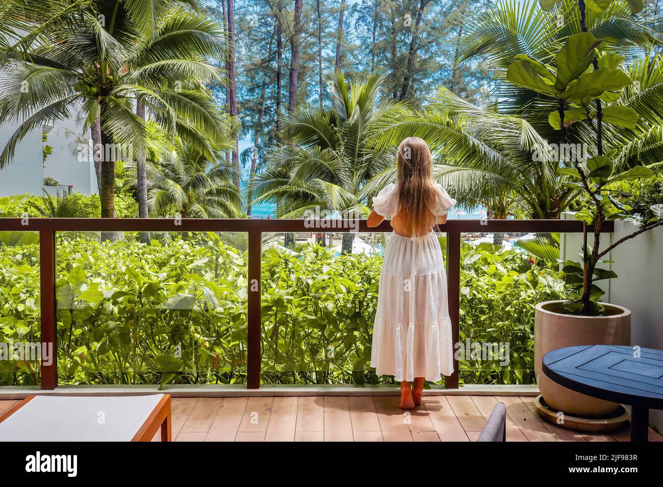 Woman standing on balcony with tropical view. Back view of female traveler relax in modern hotel room terrace with sea and palm trees view. Asia Stock Photo