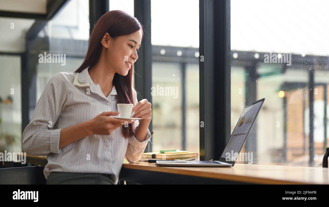 Relaxed young employee holding coffee cup and looking at screen of