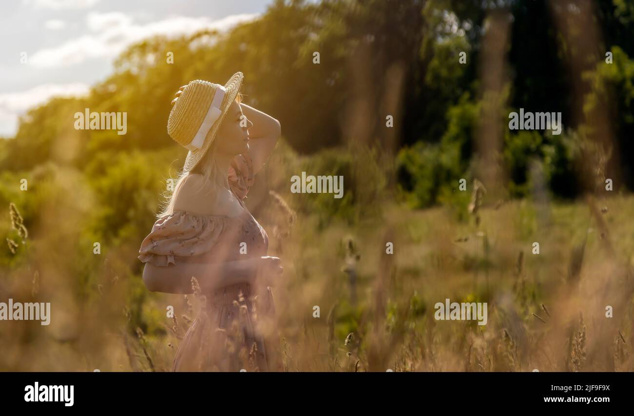 A young charming woman in a dress and a straw hat poses at sunset on a meadow. Woman in focus. Stock Photo