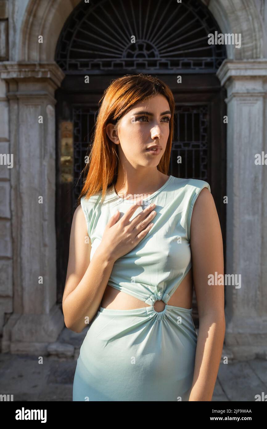 redhead woman in trendy dress touching chest and looking away on street in Venice Stock Photo