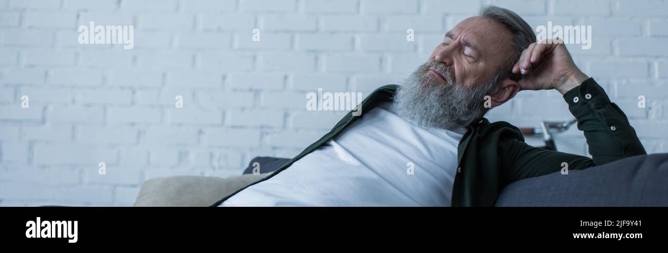 upset senior man with beard suffering crisis while sitting on couch in ...