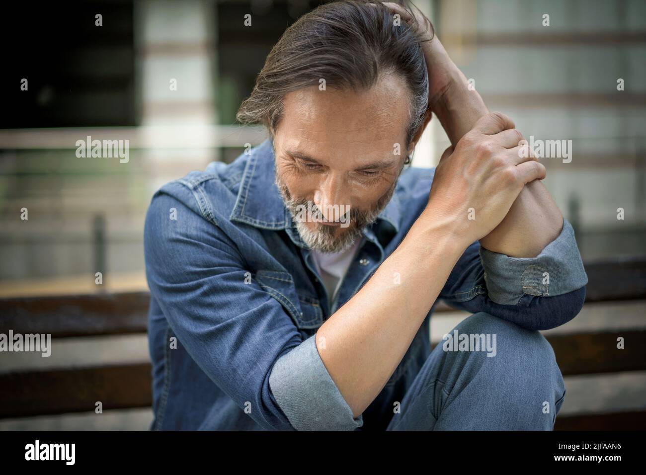 Mature grey bearded man deep in his thoughts with positive emotions on his face. Happy thinking of life middle aged man leaned his hands on knee looking down his head sitting on the bench in old town. Stock Photo