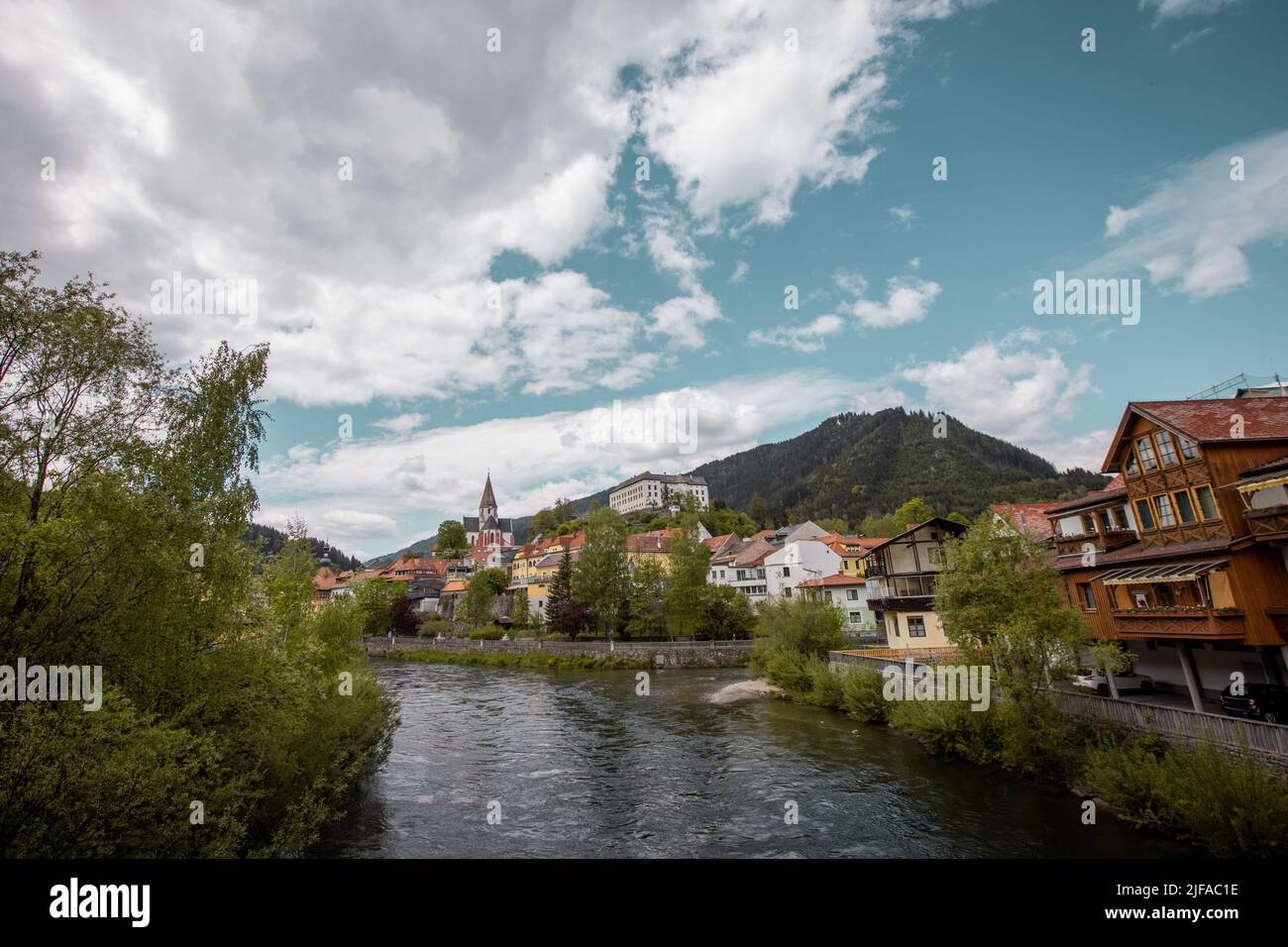 Mura or Mur river flowing through Murau city centre, in central part of austria on a cloudy but sunny day. Church and classical buildings are seen in Stock Photo
