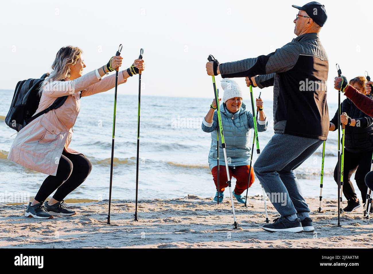 Blond woman coach teaching Scandinavian walking with sticks at sea beach coast. Exercises education for senior people, Stock Photo