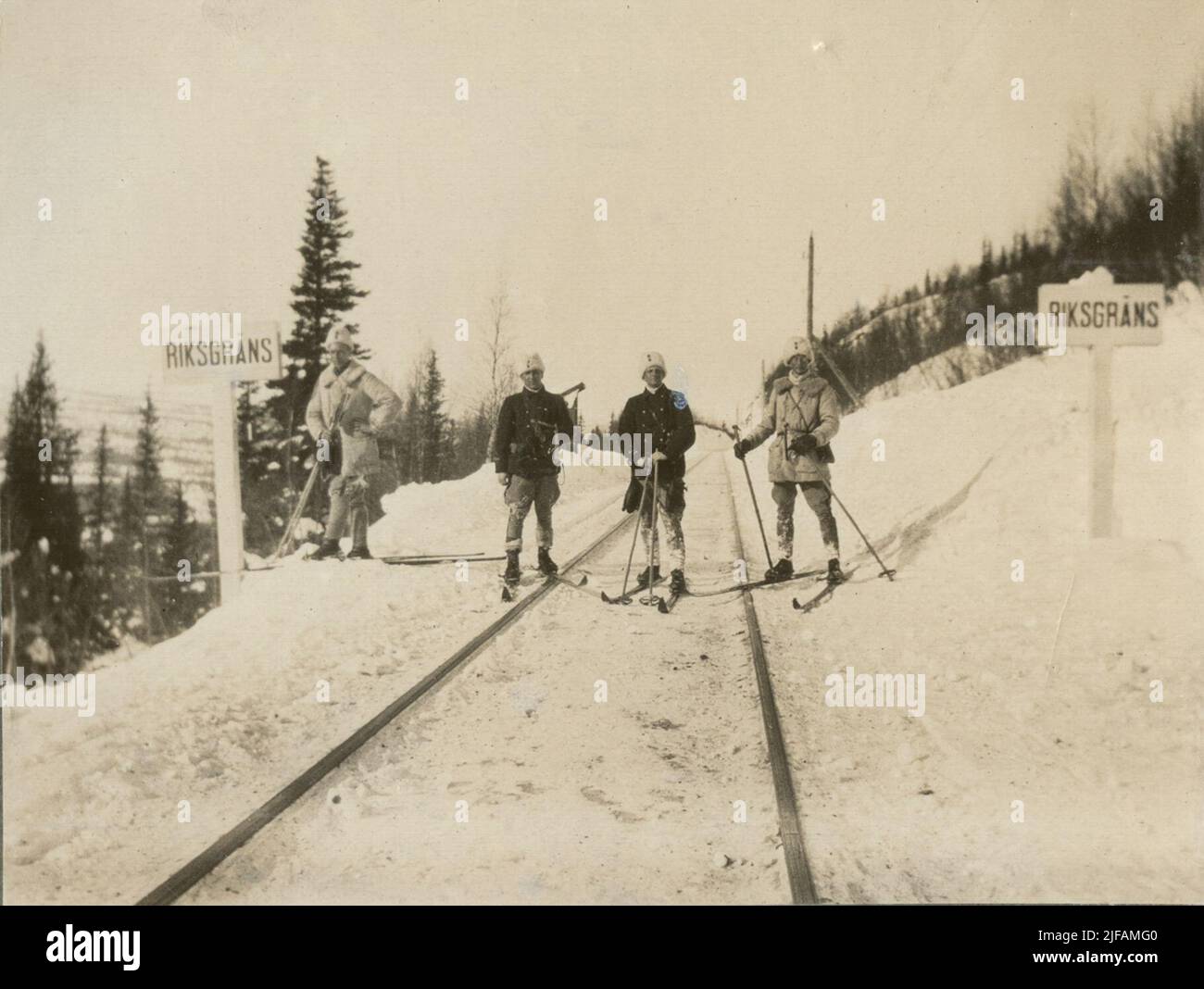 The national border between Sweden and Norway. Four soldiers on skis are on the rail rails between two signs with text 'Riksgräns. Behind the group are some trees and mountains. Stock Photo