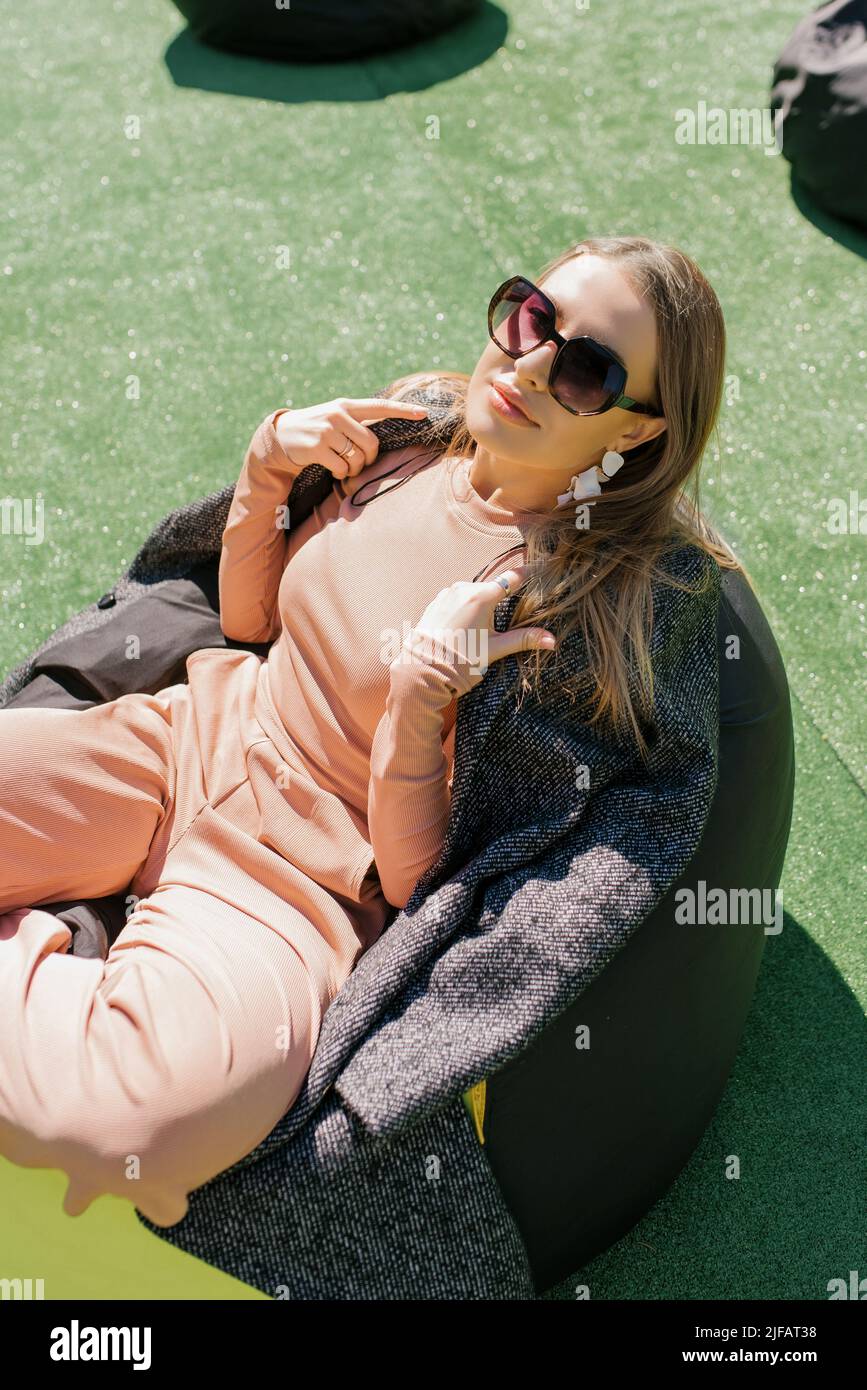 A young woman is sitting on an easy chair outdoors and enjoying a sunny day. A beautiful contented girl in sunglasses. Stock Photo