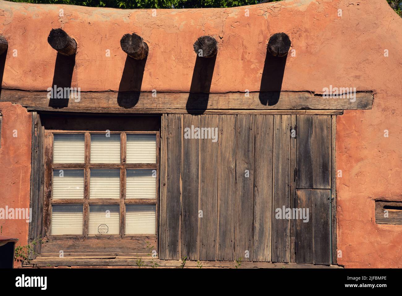 Exterior reddish brown adobe wall of a territorial style house, with vigas protruding and wooden frame paned windows in Santa Fe, New Mexico. Stock Photo