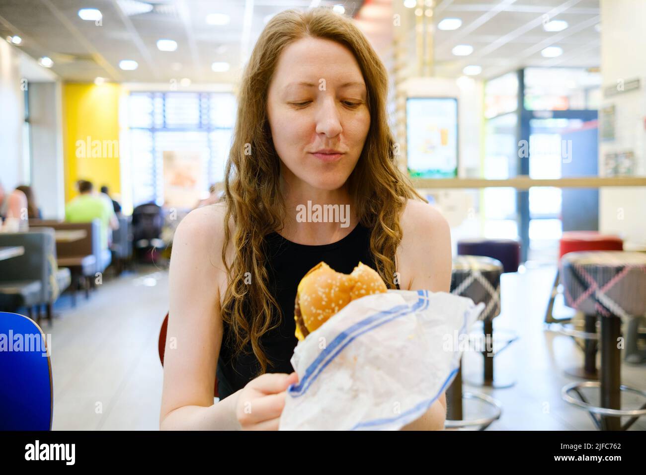 Woman with burger in a fast food restaurant. Woman opening wrapping paper, holding, biting the burger and savouring food. Fast snack on the go Stock Photo