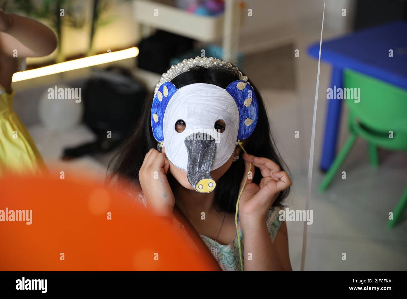 A little child girl wearing a colored animal paper mask fronting her face is isolated in the classroom background with an orange balloon. elephant pap Stock Photo
