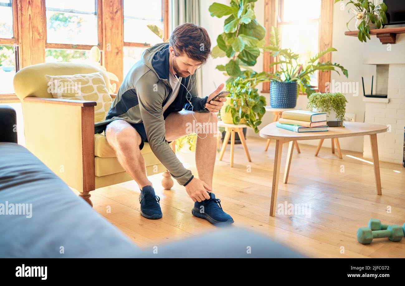 One fit young caucasian man getting dressed for exercise workout while listening to music with earphones on a cellphone at home. Guy sitting on a Stock Photo