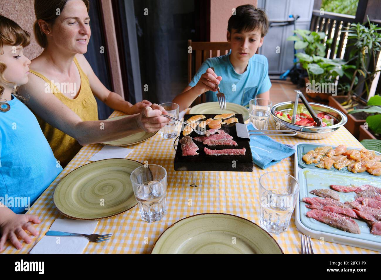 Family having lunch on the terrace of their house, the woman cooks the meat on a hot stone. Stock Photo