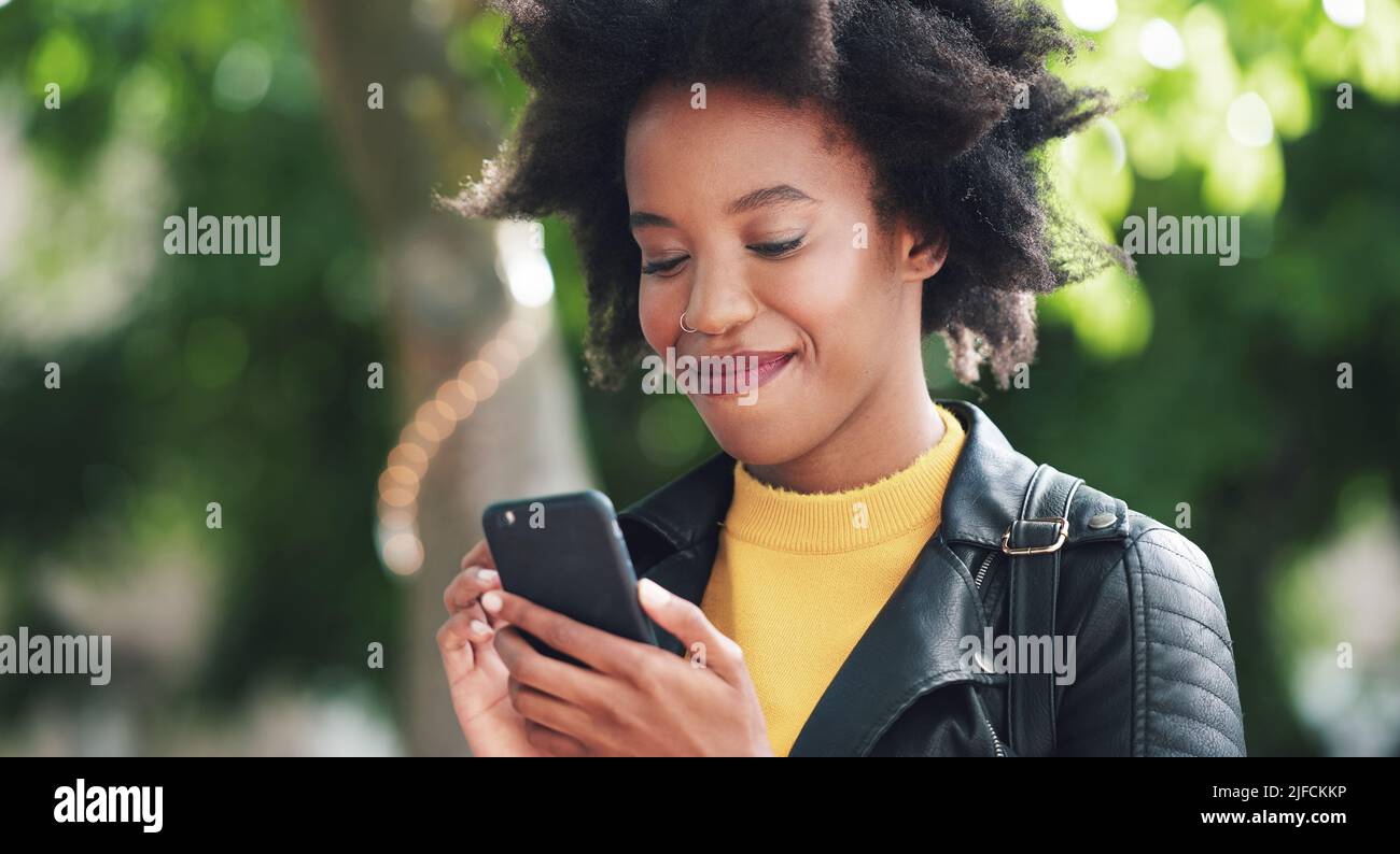 A young woman with afro using her cellphone to read text messages while in a park. A young woman in the park smiling and scrolling through apps on her Stock Photo