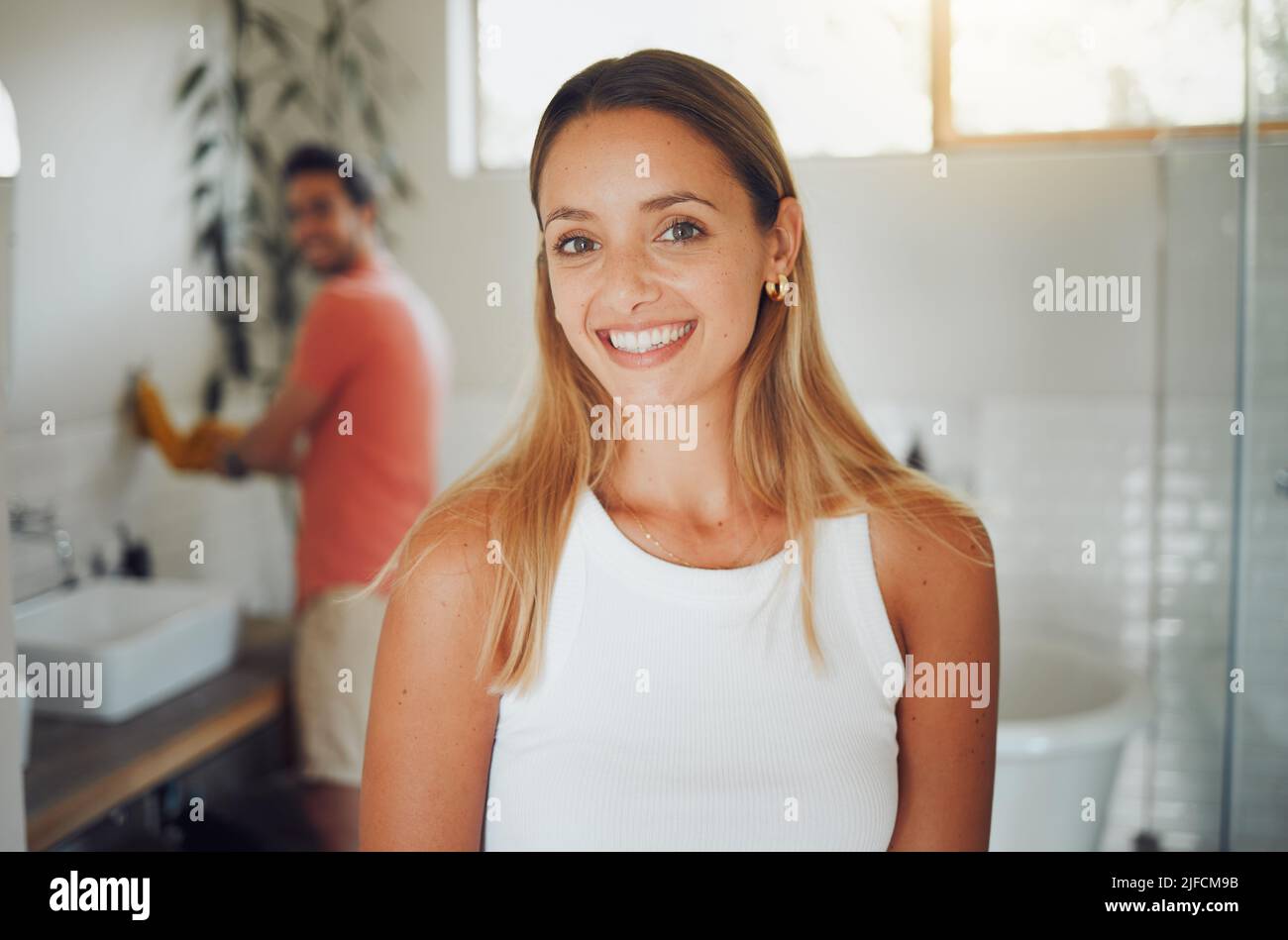 Young caucasian couple sharing a bathroom with focus on happy young woman smiling while looking at the camera in the foreground while waiting to get Stock Photo