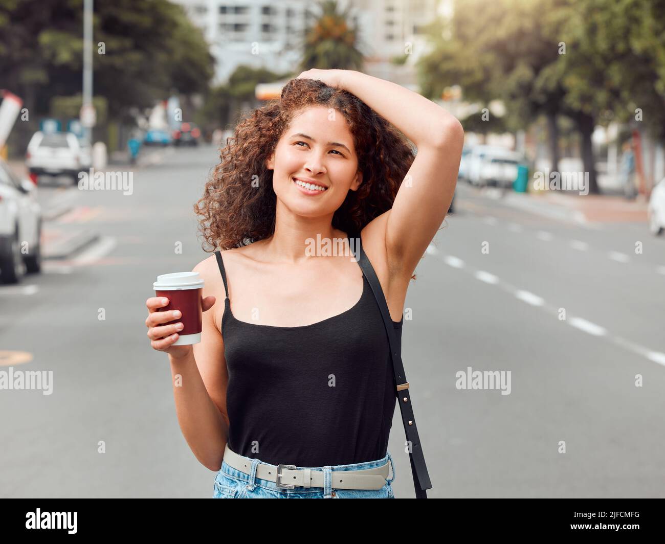 One happy beautiful young mixed race woman exploring the city while holding a takeaway coffee and touching her curly brunette hair. Hispanic Stock Photo