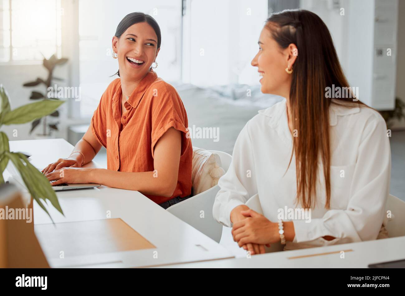 Cheerful young hispanic business woman working on a laptop while talking to a colleague in an office. Two happy coworkers working together in a Stock Photo
