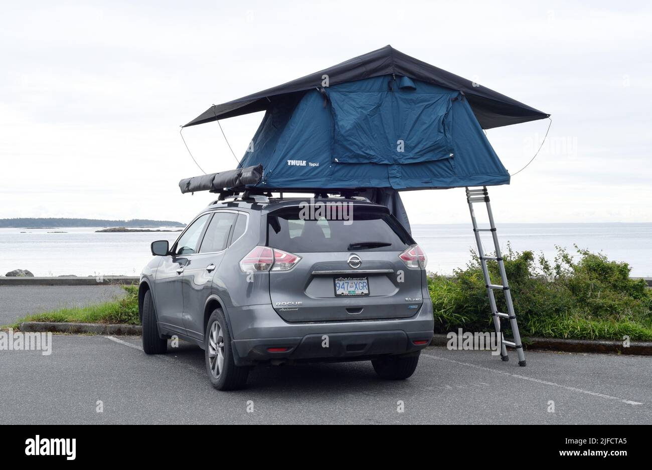 A rooftop tent is set up on top of a car parked in a waterfront parking