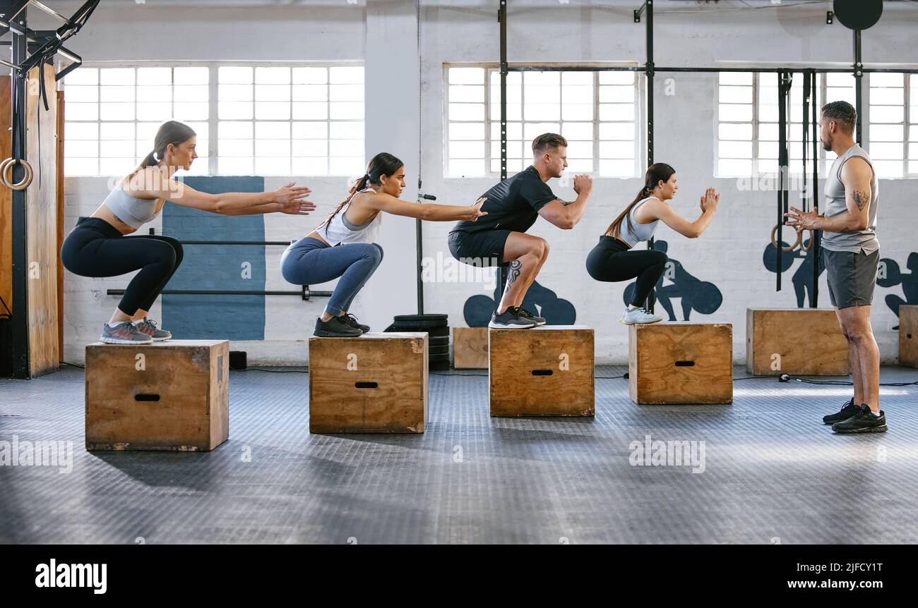 Diverse group of active young people doing box jump exercises together with a trainer in a gym. Focused athletes landing in a squat while doing Stock Photo