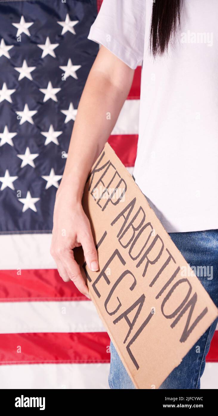 Young woman protester holds cardboard with Keep Abortion Legal sign against USA flag on background. Girl protesting against anti-abortion laws Stock Photo