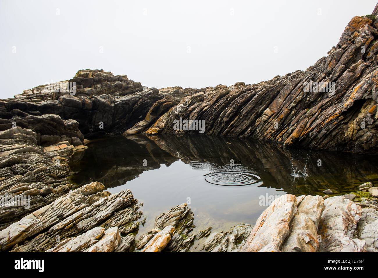 Skipping rocks on a sheltered, calm rock pool among the tilted, jagged rocks of the Tsitsikamma Coastline, South Africa. Stock Photo