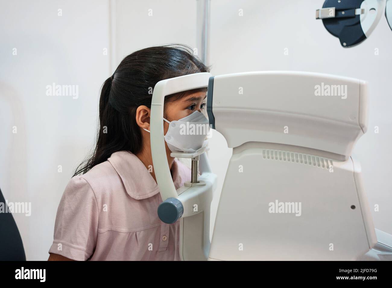 Ophthalmologist examining the eyes of an Asian girl in a clinic. They wear protective face masks. Stock Photo