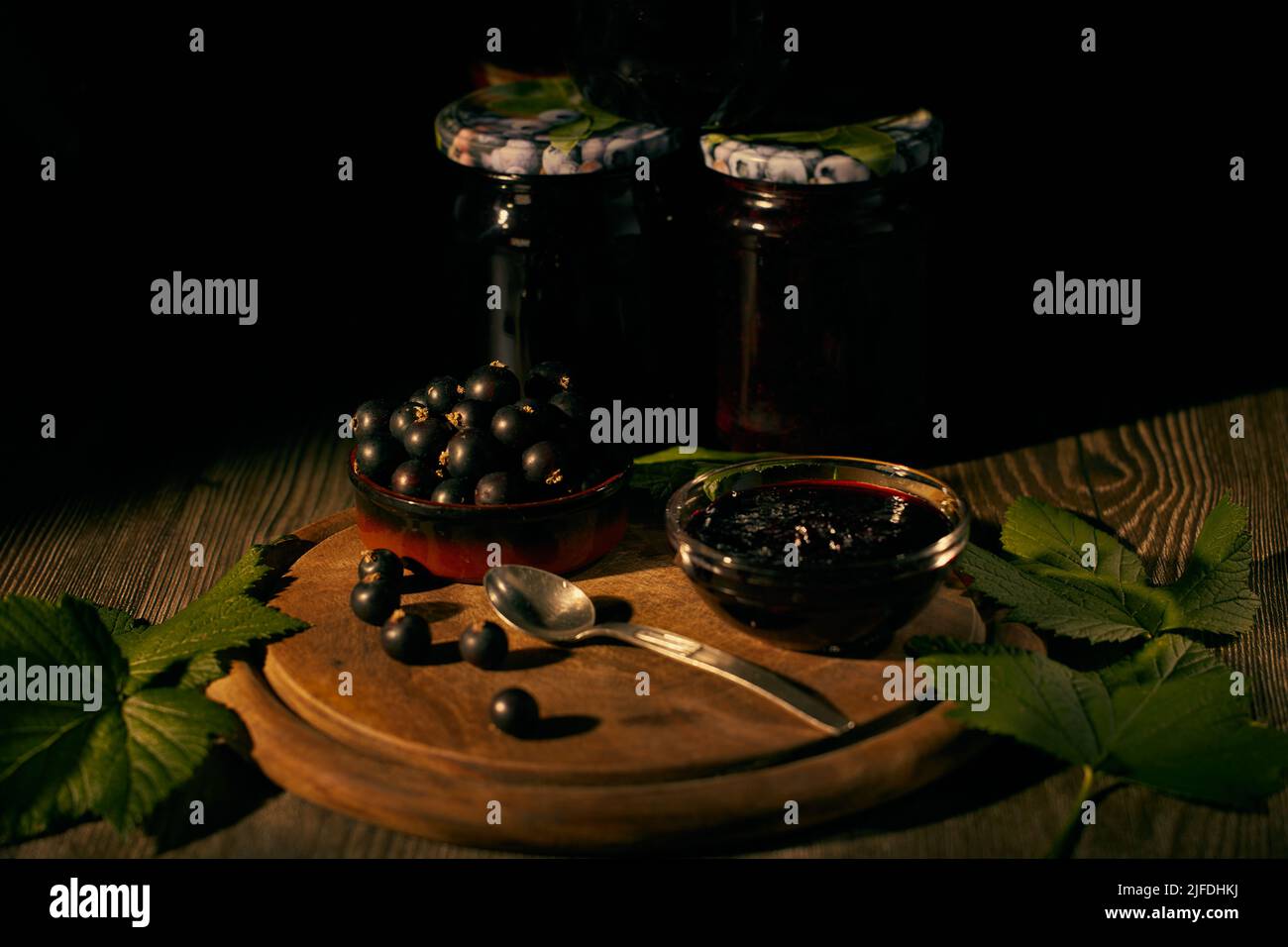 Blackcurrant in a ceramic bowl and blackcurrant jam in a glass bowl on a wooden board with green currant leaves and glass jars of jam in the backgroun Stock Photo