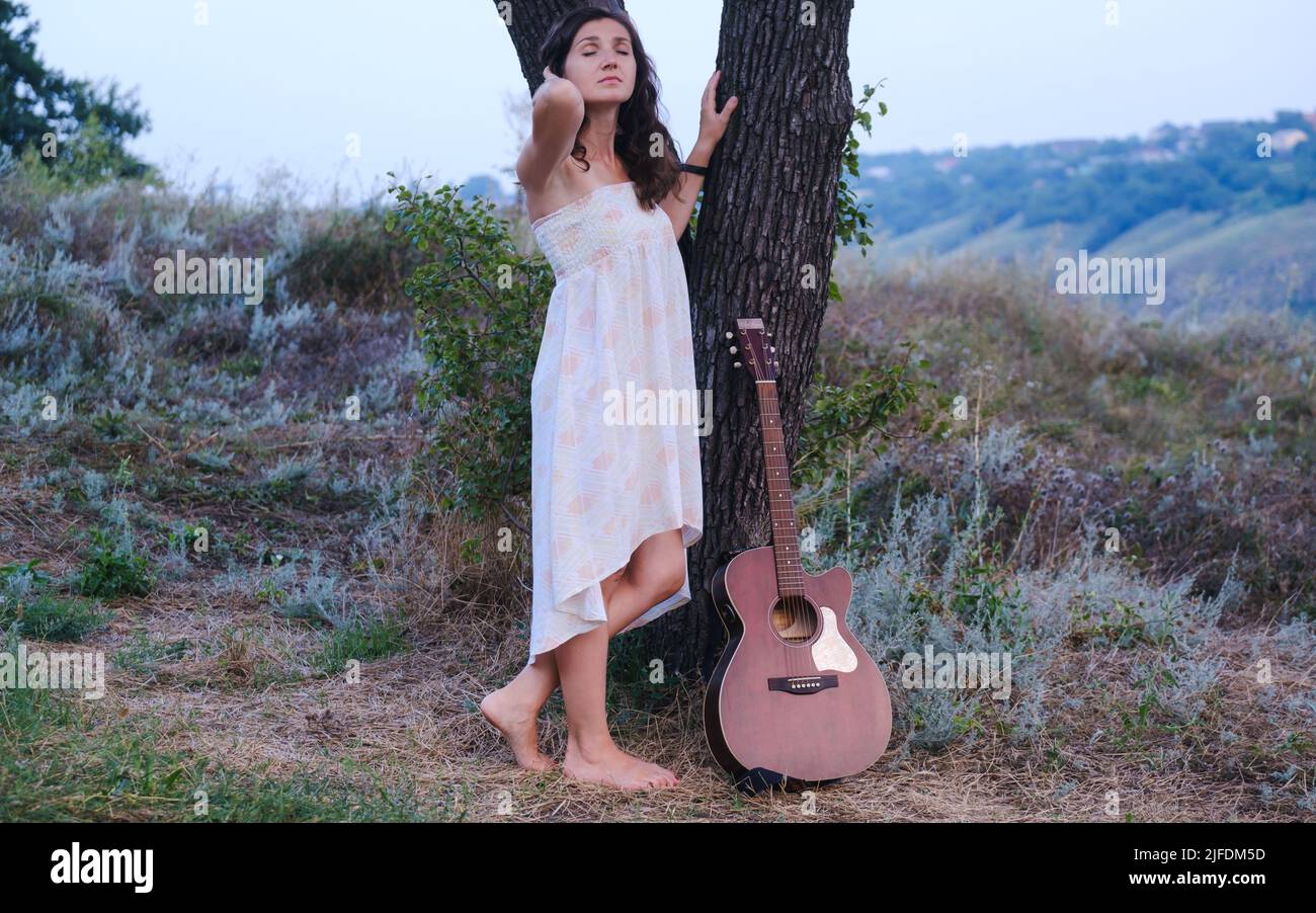 Beautiful romantic girl with dark hair and white dress with guitar outdoor. Photo of sensual woman Music background Stock Photo