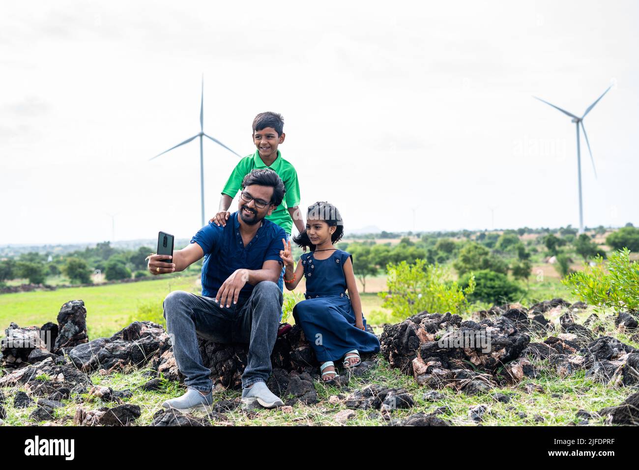 Happy father and kids taking selfie on mobile phone in front of wind turbines or power plant at hill - concept of sustainable lifestyle, technology Stock Photo