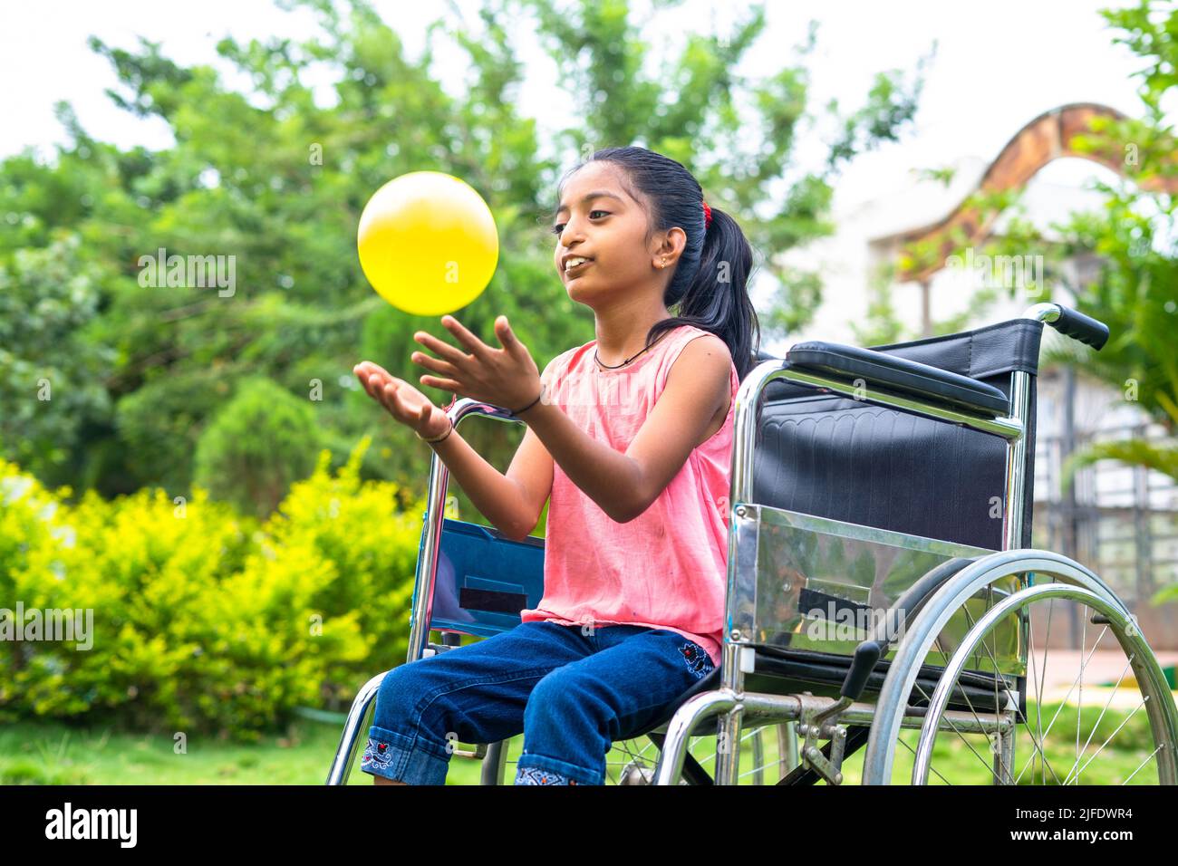 concept of happiness, freedom and enjoyment showing by Smiling alone girl kid with disability playing with ball while on wheelchair at park. Stock Photo
