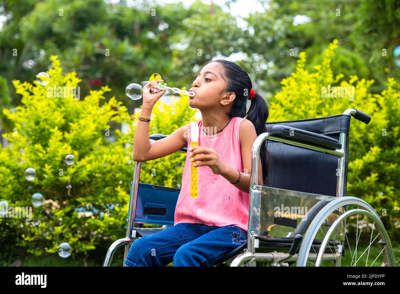 cheerful girl kid with disability playing by blowing water bubbles while using wheelchair at park - concept of playful activities, happiness and Stock Photo