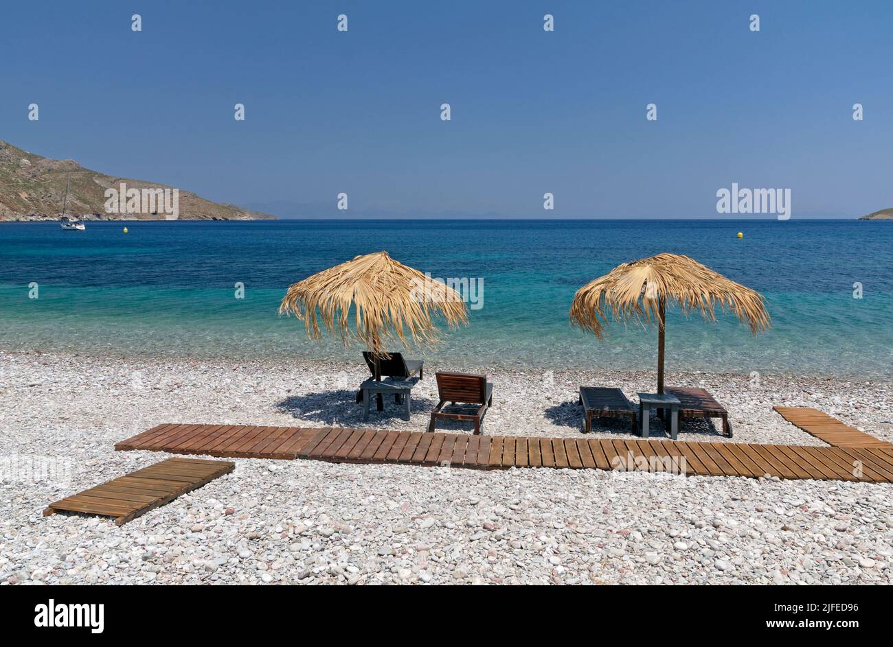 Straw beach umbrellas and bedchairs look out over idyllic beach scene with blue sky, Livadia village, Tilos island, Dodcanese, Greece Stock Photo