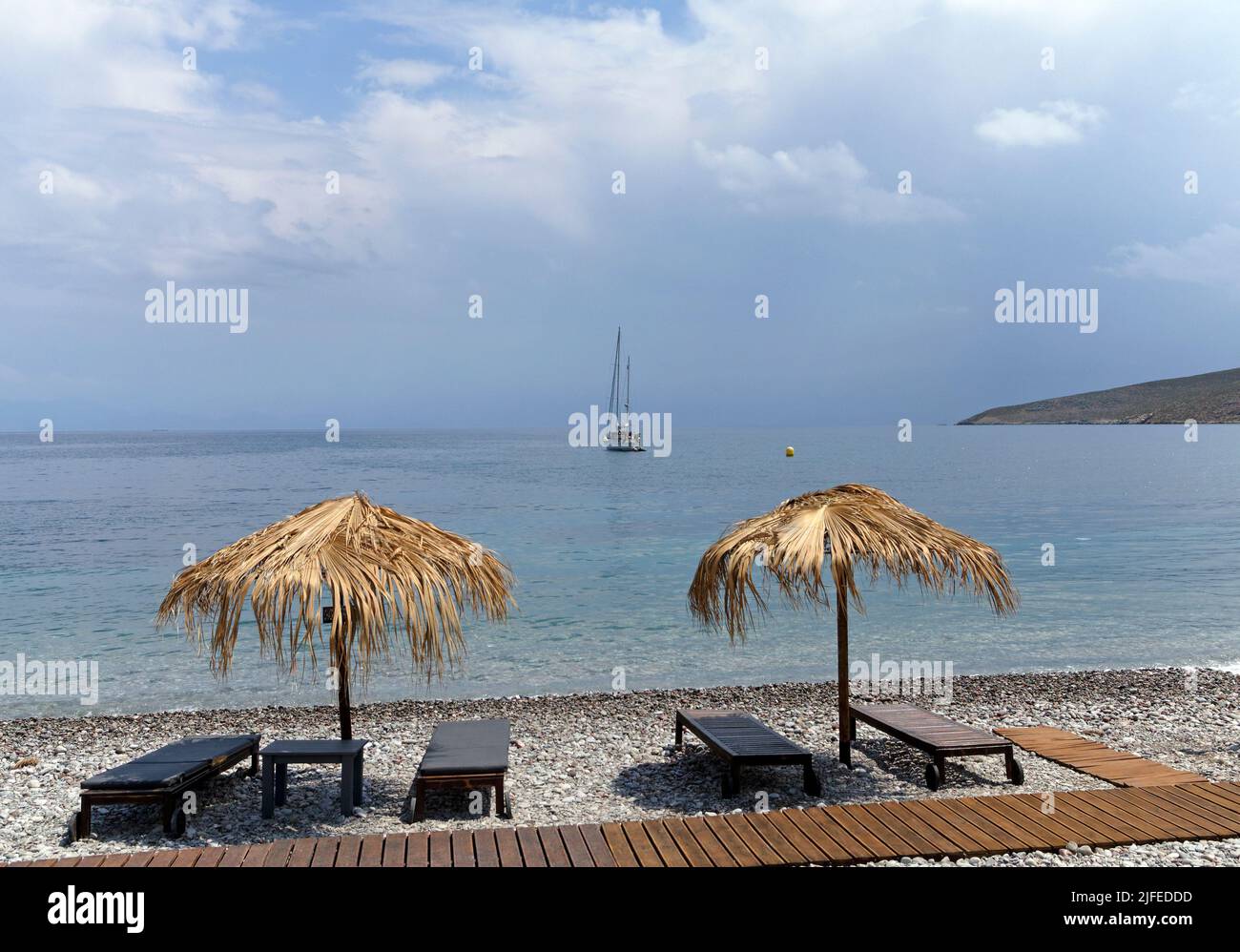 Straw beach umbrellas and bedchairs look out over idyllic beach scene with blue sky, Livadia village, Tilos island, Dodcanese, Greece Stock Photo