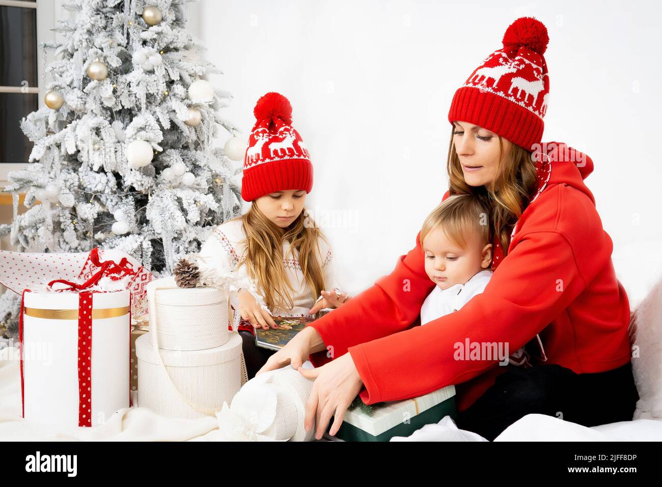Mom and children in red sweaters and hats are enjoying Christmas together. Holiday fun with the whole family around the Christmas tree. Front view. Stock Photo