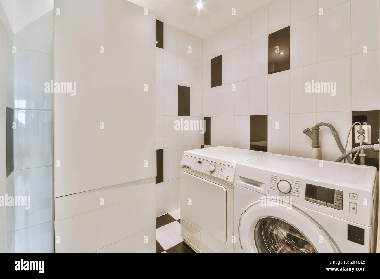 Washing machine and dryer in a bright room with white and black tiles and white pipes on the wall Stock Photo