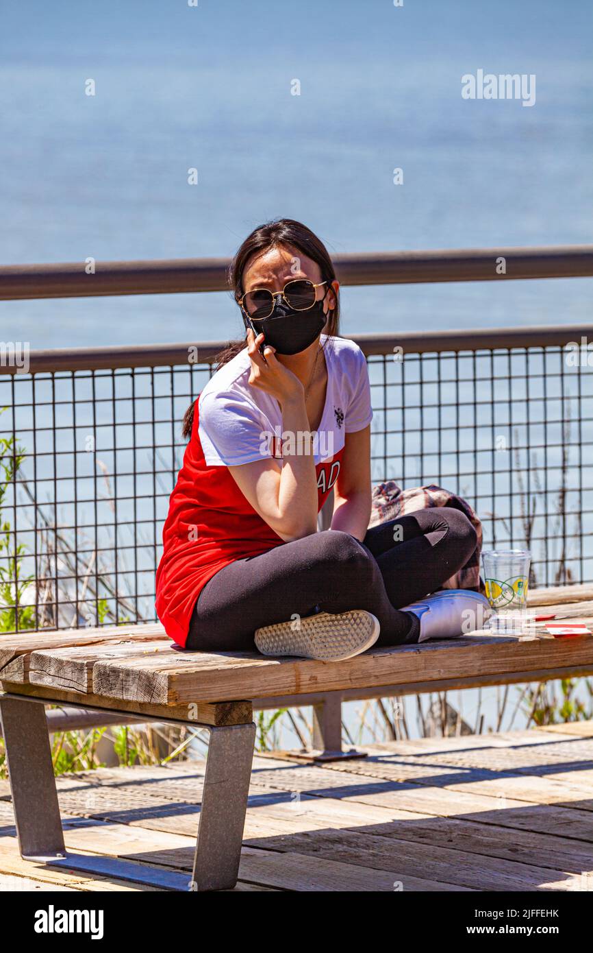 Young woman talking on her phone wearing a black Covid face mask along the Steveston waterfront on Canada Day 2022 Stock Photo